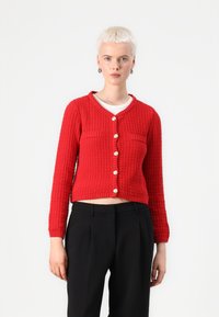 Cardigan en laine rouge avec un motif texturé, dotée d'un col en V, de poches avant et de sept boutons. Longueur courte, associée à un pantalon noir.