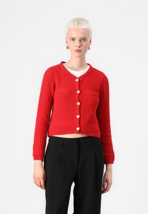 Rode gebreide cardigan met een textuurpatroon, voorzien van een v-hals, steekzakken en zeven knopen. Verkorte lengte, gecombineerd met zwarte broeken.