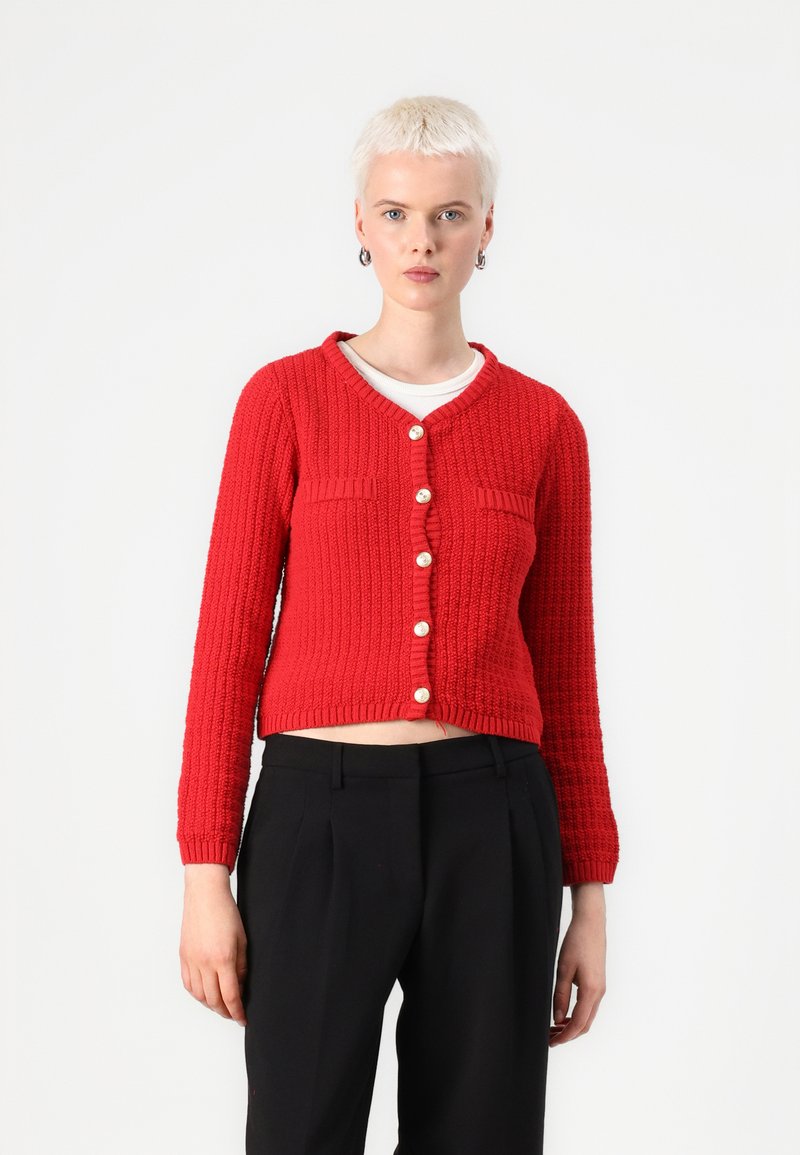 Cardigan en laine rouge avec un motif texturé, dotée d'un col en V, de poches avant et de sept boutons. Longueur courte, associée à un pantalon noir.