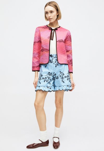 Η γυναίκα φοράει ροζ υφασμάτινο τζάκετ πάνω από λευκή μπλούζα, συνδυασμένο με ανοιχτό μπλε κεντημένα σορτς με βολάν και καφέ παπούτσια Mary Jane με λευκές κάλτσες.