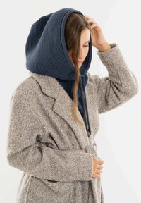 Gebreide marineblauwe capuchon met ribbeltextuur en een grijze wollen jas. De jas heeft een taille met riem en brede mouwen, waardoor er een gelaagde look ontstaat.