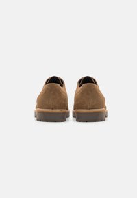 Scarpe in suede marrone con punta arrotondata, suola in gomma a contrasto e un tacco piccolo. La parte posteriore presenta linee morbide e pulite con dettagli minimi.