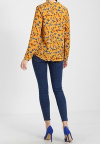 Mujer vistiendo una blusa larga de manga larga florada en color mostaza, jeans ajustados oscuros y zapatos de tacón alto azules brillantes, de pie con la espalda visible.