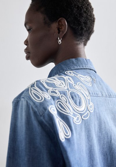 Camicia in denim con design ricamato bianco sulla schiena, colletto corto e tessuto morbido. Modello che mostra il profilo laterale, con orecchini a cerchio.