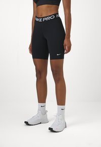 Shorts Nike Pro noirs avec une taille haute, arborant un logo blanc et une texture lisse et élégante. Associés à des chaussures et des chaussettes de sport blanches.