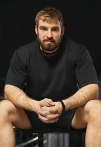 Zwarte sportieve T-shirt met een ronde hals en logo-detail. Het model heeft kort haar en een baard, en zit met gekruiste handen op de knieën.