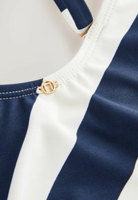 Marineblauwe en witte gestreepte stof met een gladde textuur. Bevat een gouden hardware-detail nabij de naad, wat het ontwerp versterkt.