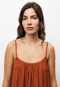 Rostfarbendes, ärmelloses Kleid mit dünnen Trägern, das einen gerafften Ausschnitt und einen weichen, fließenden Stoff aufweist. Glatte Textur, lockere Passform.