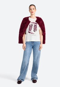 Mujer vistiendo un abrigo burdeos de felpa sobre una camiseta blanca con un gráfico decorado con botas vaqueras moradas, combinado con unos pantalones de mezclilla de pierna ancha azul claro y zapatillas negras.