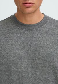 Maglione grigio a girocollo con colletto a costine e texture liscia, che mette in evidenza la finitura morbida del tessuto e le sottili variazioni di colore.