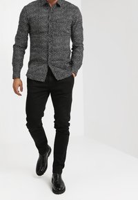 Camisa negra de manga larga con lunares blancos, con botones en el frente, combinada con pantalones negros ajustados y zapatos negros brillantes.