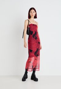 Vestido longo floral vermelho com alças finas, sobreposição transparente e detalhes em preto. Combinado com botas pretas de salto grosso e uma pequena bolsa preta.