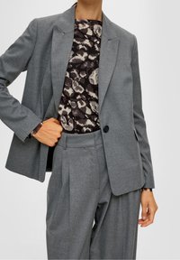Šedý střižený blejzr s výpustkovými klopy, jedním knoflíkem a postranními kapsami, nošený přes vzorovaný top. Hladká textilie, strukturovaná silueta.