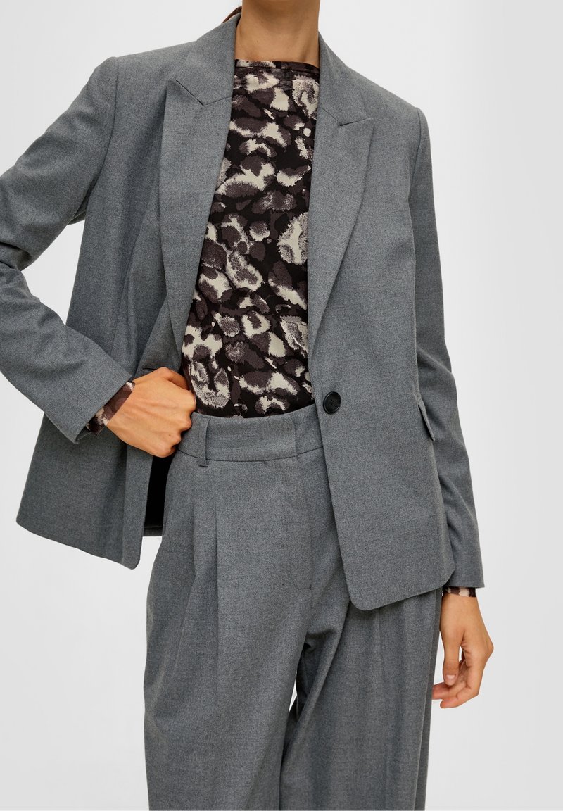 Šedý střižený blejzr s výpustkovými klopy, jedním knoflíkem a postranními kapsami, nošený přes vzorovaný top. Hladká textilie, strukturovaná silueta.
