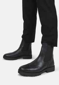 Bottines en cuir noir avec panneaux latéraux élastiques, détails de brogue texturés et une semelle en caoutchouc robuste pour une bonne adhérence et durabilité.