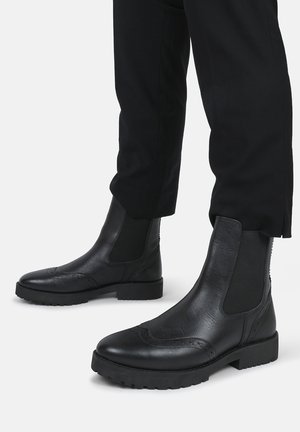 Stivali alla caviglia in pelle nera con pannelli laterali elasticizzati, dettagli brogue testurizzati e una suola in gomma resistente per aderenza e durabilità.