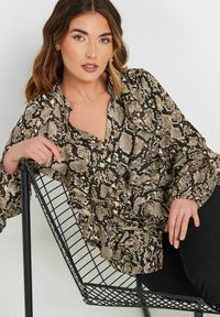 Braun- und schwarz gemusterte Schlange-Bluse mit Rüschendetails, V-Ausschnitt und langen Ärmeln, getragen über schwarzen Hosen, posiert auf einem Metallstuhl.