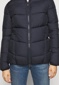 Doudoune bleu marine avec fermeture éclair à l'avant, dotée de sections matelassées et de poignets élastiques. Tissu lisse avec un léger éclat.