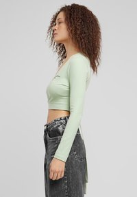 Világoszöld, hosszú ujjú crop top V-nyakkal és fekete hímzett részlettel, magas derekú fekete farmernadrággal párosítva.