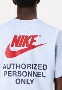 Gaiši zilas kokvilnas T-krekls ar izteiktu sarkano Nike logo un tekstu "TIKAI AUTORIZĒTIEM DARBINIEKOM" melnā krāsā uz aizmugures. Īsas piedurknes.