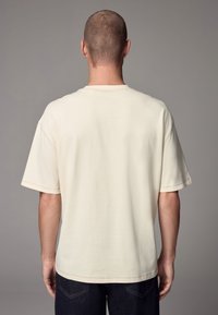 Cream-colored, short-sleeve t-shirt with a relaxed fit and a round neckline. Features visible stitching and smooth texture. Worn with dark jeans.