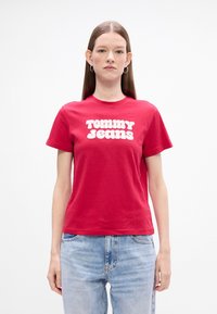 Punane puuvillane T-särk, millel ees on julge valge tekst "TOMMY JEANS", bulid ümmargune kaelus ja lühikesed varrukad. Kandmisel heledate siniste teksadega.