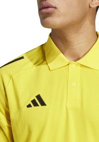 Gele polo gemaakt van gestructureerde stof met een tweeknoopsluiting, voorzien van zwarte strepen op de schouders en een zwart logo op de borst.