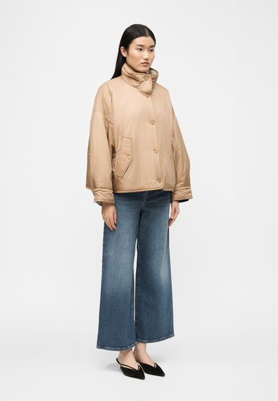 Νεαρή γυναίκα με μακριά μαύρα μαλλιά, που φοράει μπεζ μπουφάν με επένδυση, μπλε τζιν παντελόνι με φαρδιές παντελόνες και μαύρα slingback παπούτσια με μυτερή μύτη, στέκεται μπροστά σε λευκό φόντο.