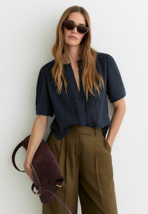 Žena s dlhými vlnitými vlasmi v čiernej plisovanej blúzke, hnedých nohaviciach s vysokým pásom, slnečných okuliaroch a so svojou veľkou tmavofialovou kabelkou.