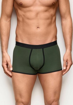 Hombre sin camiseta de pie contra una pared blanca, vistiendo calzoncillos bóxer verdes oscuros con ribete y cintura negros.