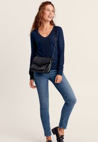 Schwarze Umhängetasche mit strukturiertem Äußeren, Kettenriemen und schrägem Klappenverschluss. Getragen mit einem marineblauen Pullover und blauen Jeans, was ein lässiges Outfit zeigt.