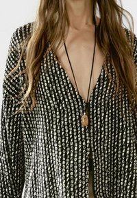 Kobieta nosi czarny i biały, teksturowany kardigan zapinany z przodu oraz naszyjnik z zawieszką z brązowym kamieniem na czarnym sznurku.