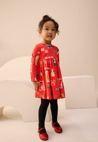 Rode langemouwen jurk met speelse kerstpatronen, met een gather in de taille en een gegolfde zoom. Gecombineerd met zwarte panty’s en rode pailletten schoenen.