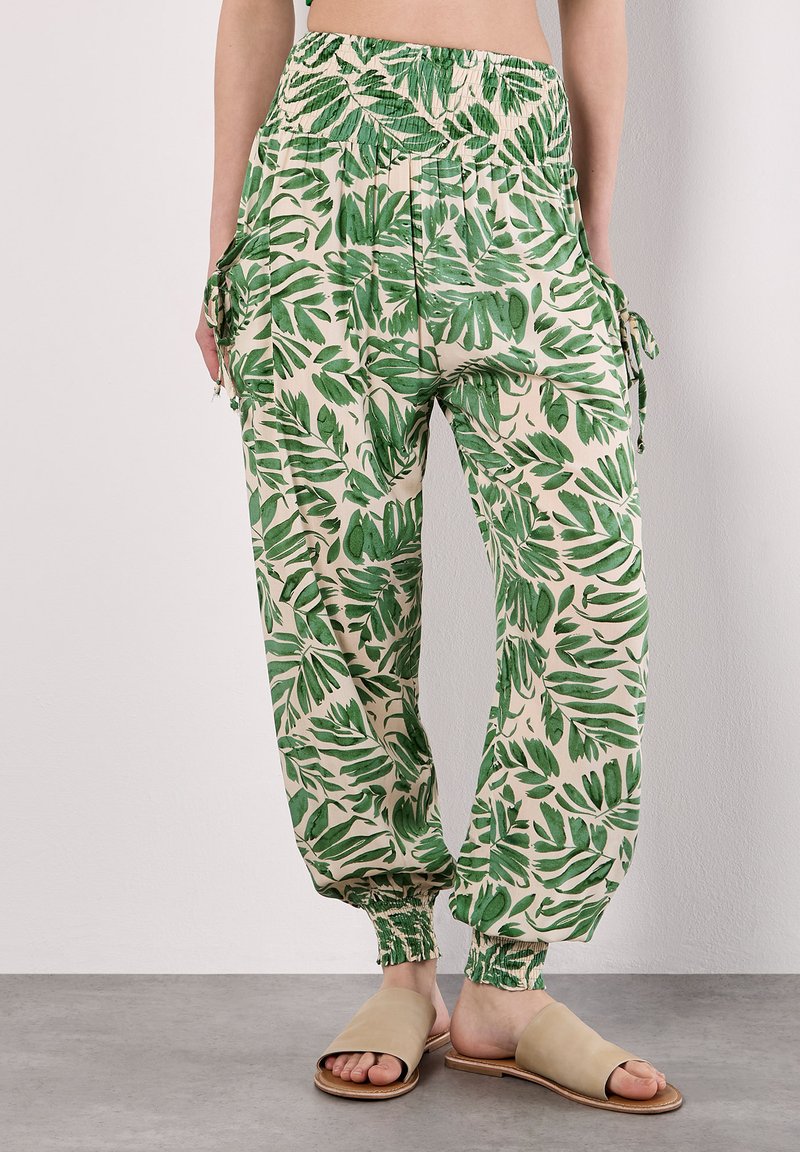 Une personne portant un pantalon à motif de feuilles avec une taille élastique se tient sur une surface gris clair, complété par des sandales à glissière beiges.