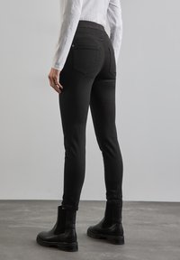 Zwarte stretchy skinny jeans met een hoge tailleband, met achterzakken, gecombineerd met zwarte enkelboots, op een grijze betonnen vloer.