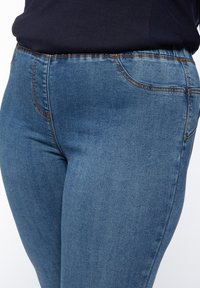 Blå denimjeans med en slät yta, som har fem fickor, kontrasterande sömmar och ett elastiskt midjeband för ökad komfort.