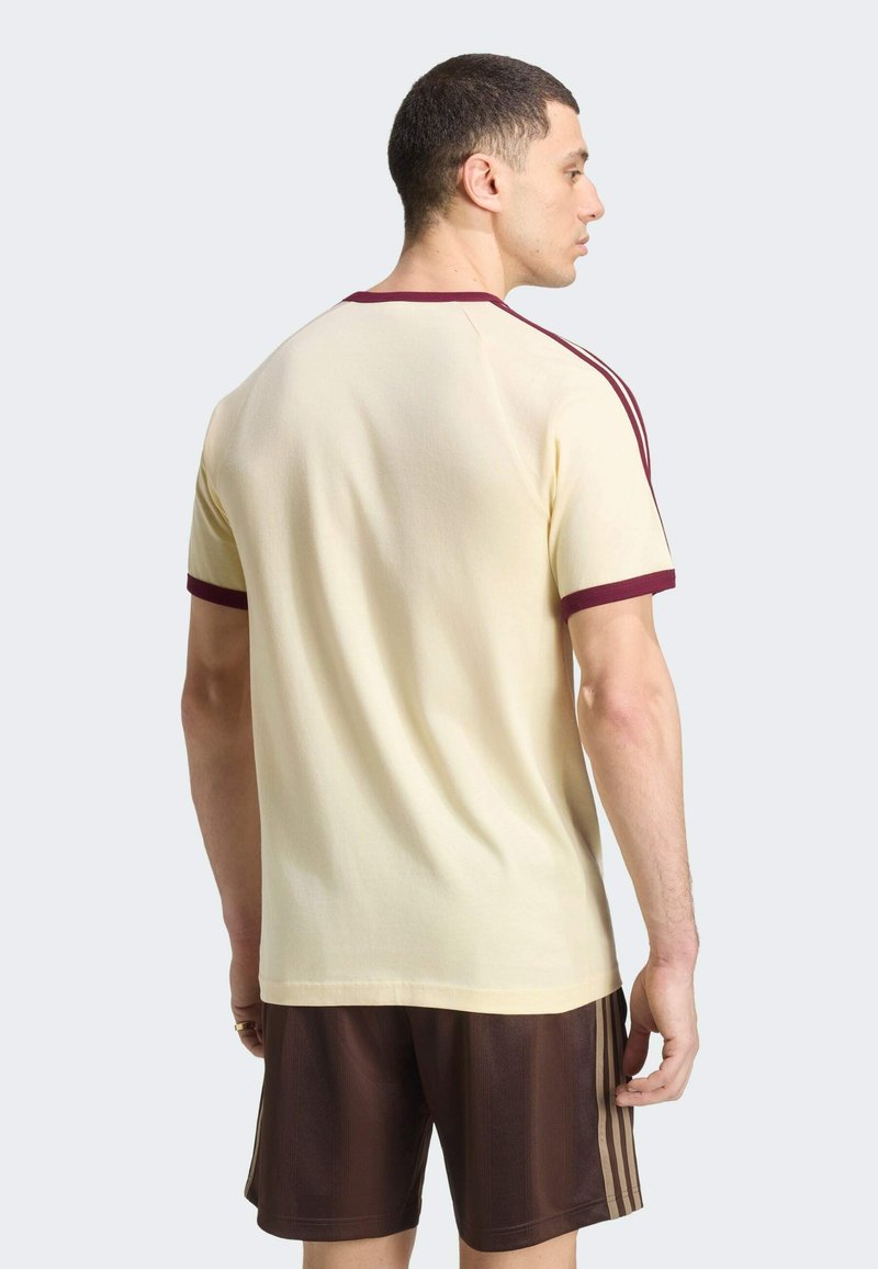 Hombre de espaldas, vistiendo una camiseta crema con ribete granate y pantalones cortos marrones con rayas beige en los lados, sobre un fondo liso.