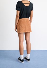 Personne portant un haut noir à manches courtes avec dos ouvert, une mini-jupe en velours côtelé marron, des chaussettes blanches et des baskets noires, debout sur un sol bleu.