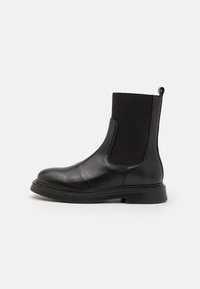 Botte de cheville en cuir noir avec panneaux latéraux élastiques, surface texturée, bout rond et semelle durable. Design minimaliste avec une languette.