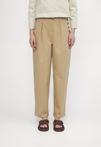 Beige bomullsbukser med folder, med sideknapper og en smal passform. Brukt med flettede brune sandaler.