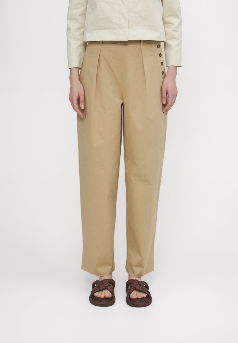 Beige bomullsbukser med folder, med sideknapper og en smal passform. Brukt med flettede brune sandaler.