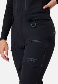 Sorte vandtætte overalls lavet af slidstærkt stof, med lynlås lommer, justerbare stropper og et logo på låret.