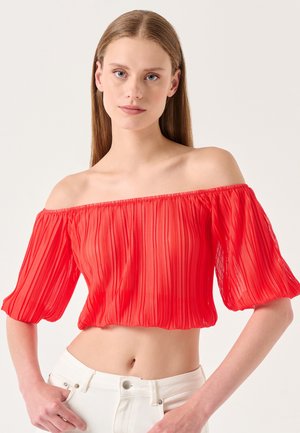 Crop top rosso, scollato sulle spalle, con pieghe e maniche a palloncino. Il tessuto è traslucido e morbido, abbinato a jeans bianchi a vita alta.