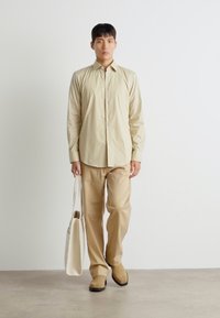 Cămașă bej cu nasturi, cu mâneci lungi, asortată cu pantaloni și pantofi de aceeași culoare. O geantă albă este ținută într-o mână. Design simplu și curat.