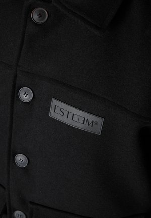 Black wool coat with black buttons and a rectangular patch labeled "ESTEEM" on the front chest pocket.