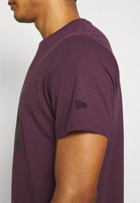 Lila bomulls t-shirt med korta ärmar, rund halsringning och en svart broderad logotyp på vänster ärm. Slät textur.