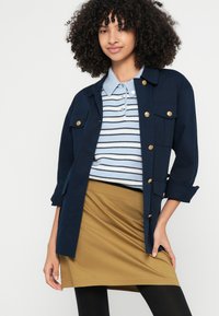 Μπλε ναυτικό βαμβακερό σακάκι με χρυσά κουμπιά πάνω από ανοιχτό μπλε και λευκό ριγέ πόλο. Φούστα σε ελιά πράσινο, μαύρο καλσόν και σγουρά μαλλιά.