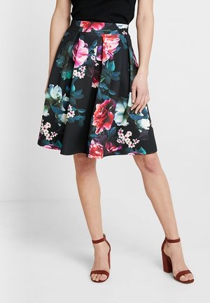 Jupe noire évasée avec un grand imprimé floral coloré portée avec un haut noir et des sandales marron à talons hauts et bride cheville.