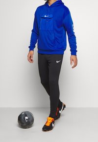 Sweatshirt bleu avec poche avant, pantalon de sport noir ajusté, chaussures de football noires avec des accents orange, et un ballon de football noir au sol.