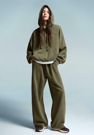 Hoodie surdimensionné vert olive avec cordon de serrage et poche frontale, associé à un pantalon de jogging large assorti et des baskets marron.